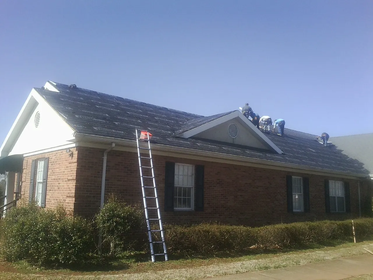Crew tearing shingles off a brick ranch roof with a ladder set — Metal Roof Repair work by our crew in Rosman