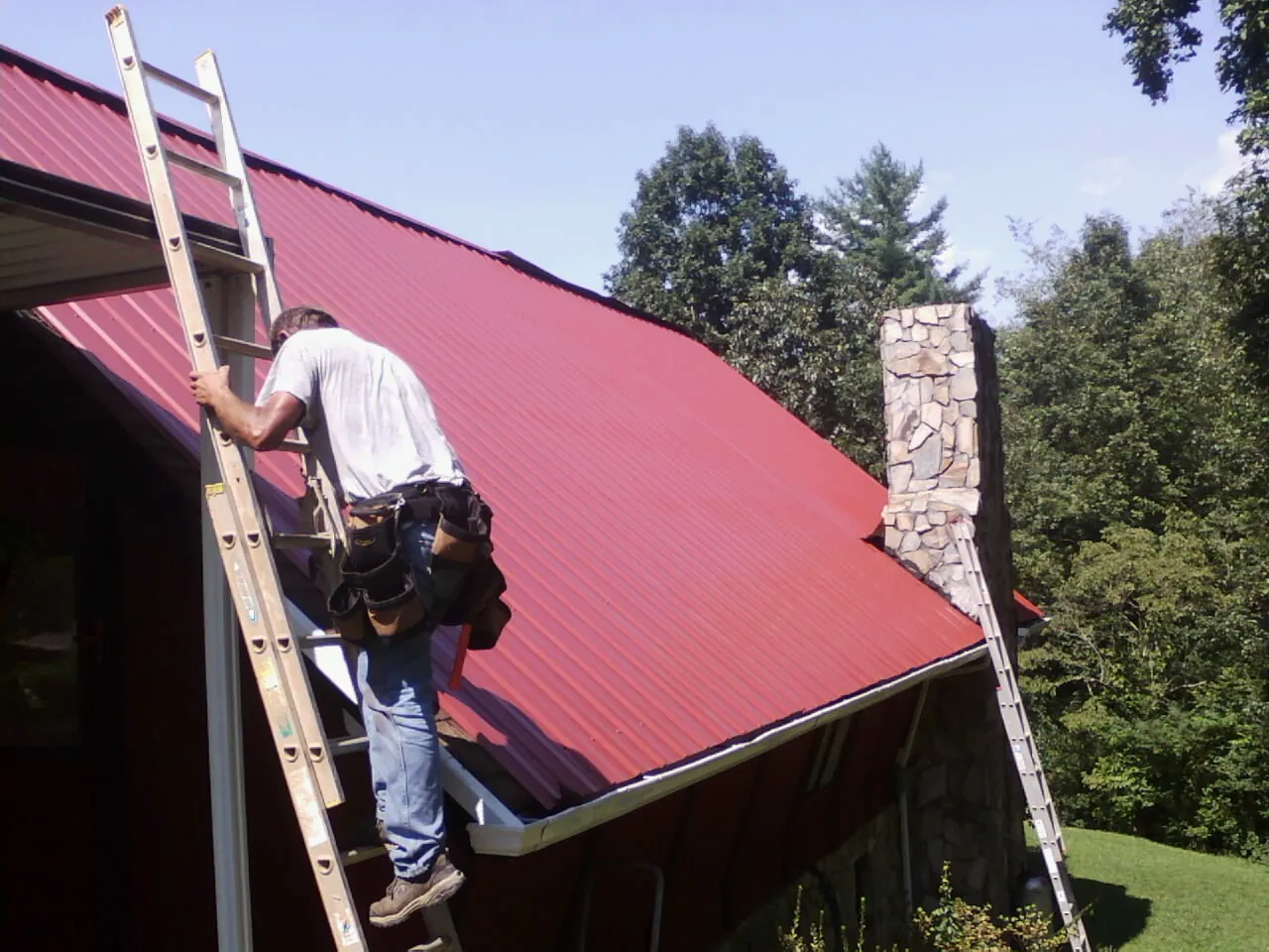 Roofer on a ladder beside a red metal roof and stone chimney — Soffit & Fascia Repair by Roofing Brothers in Rosman